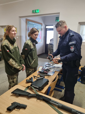 Umundurowany policjant stoi. Trzyma broń. Obok niego stoją dwie kobiety ubrane w mundur ćwiczebny.