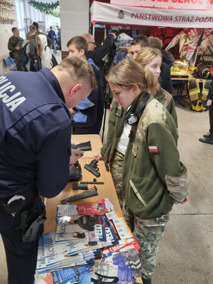 Umundurowany policjant stoi, pochyla się. Obok niego stoją ubrane w mundur ćwiczebny kobiety.