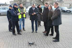 Dwóch umundurowanych funkcjonariuszy stoi. Obok stoją osoby. Przed nimi leży dron.