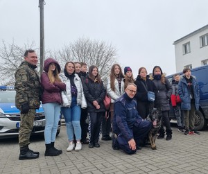 Młodzież stoi, umundurowany policjant z psem kuca.