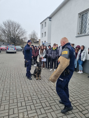 Młodzież stoi. Widać umundurowanego policjanta z rękawem do tresury psów policyjnych.