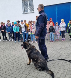 Policjant umundurowany stoi z psem służbowym. Obok stoją dzieci.