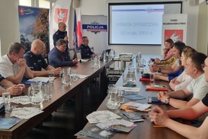 Umundurowany policjant stoi, inne osoby siedzą.