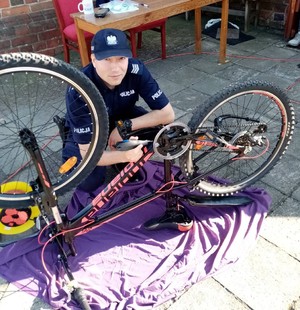 Umundurowany policjant kuca przy rowerze. W ręce trzyma urządzenie do grawerowania.