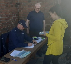 Umundurowany policjant siedzi w ręku trzyma długopis. Obok stoją dwie osoby.