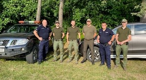 Umundurowani policjanci stoja z funkcjonariuszami Straży Leśnej i Straży Łowieckiej na tle drzew i radiowozów.