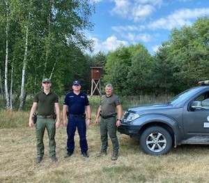 Umundurowany policjant stoi z funkcjonariuszem Straży Leśnej i Straży Łowieckiej na tle drzew.