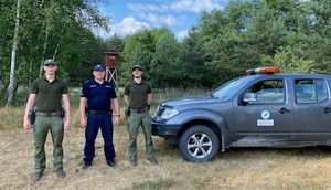 Umundurowany policjant stoi z funkcjonariuszem Straży Leśnej i Straży Łowieckiej na tle drzew.