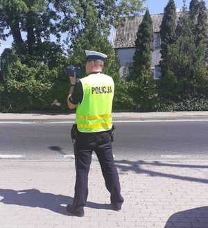 Umundurowany policjant stoi tyłem, w rękach trzyma przyrząd służący do pomiaru prędkości przez kierujących pojazdami.