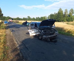 Samochód uszkodzony w wyniku zdarzenia drogowego. W oddali widać stojącego policjanta i radiowóz.